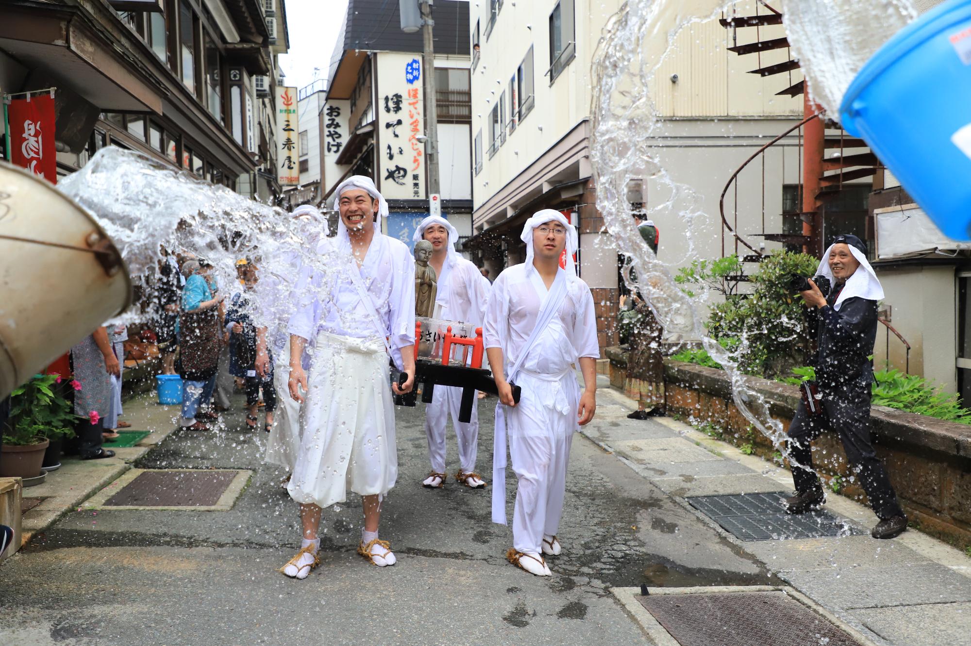 温泉街で地蔵神輿を持った白装束の姿の若者に道路の両脇から温泉のお湯がかけられている写真
