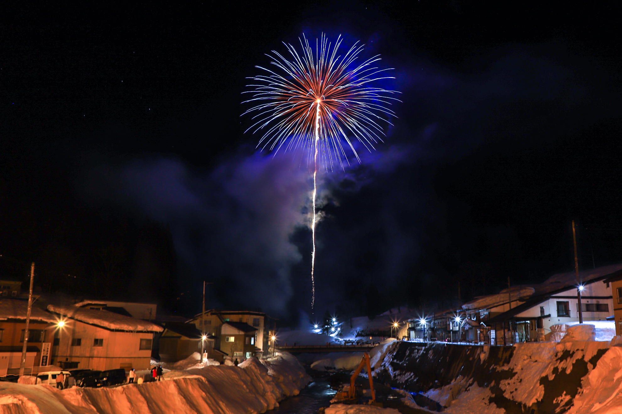 中央に川が流れ、両脇に雪が積もっている家々が立ち並んでおり、花火が打ち上げられた写真。