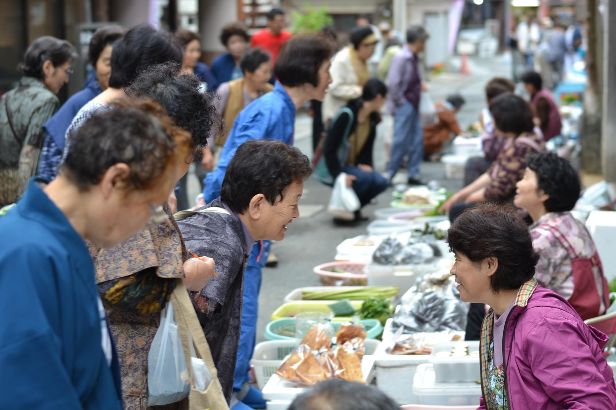 肘折温泉の旅館の軒先に山菜や野菜などがずらりとならび、浴衣姿の当時客が買い物を楽しむ写真