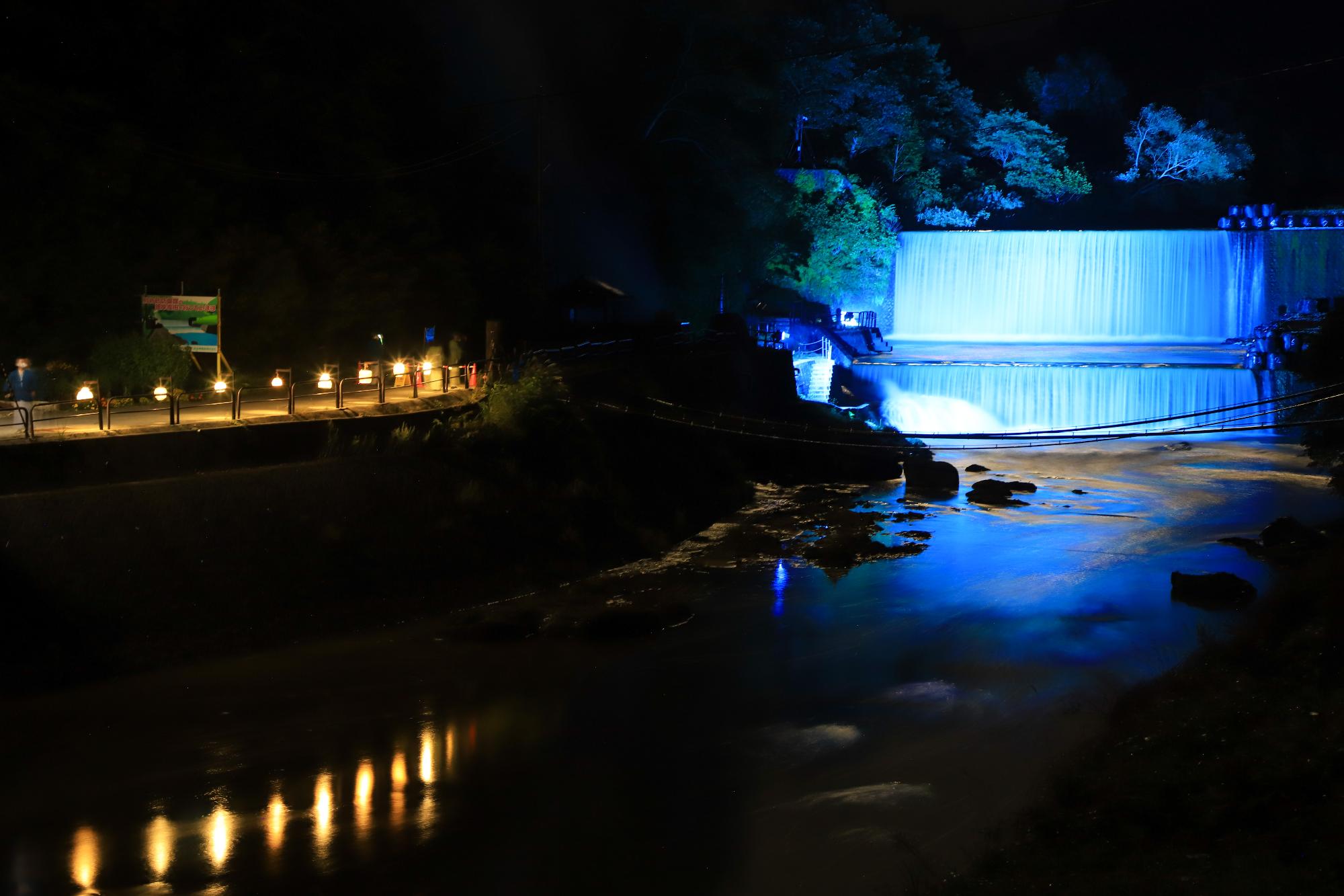 ダムが青色にライトアップされ、通路には黄色の光で道しるべになっている写真