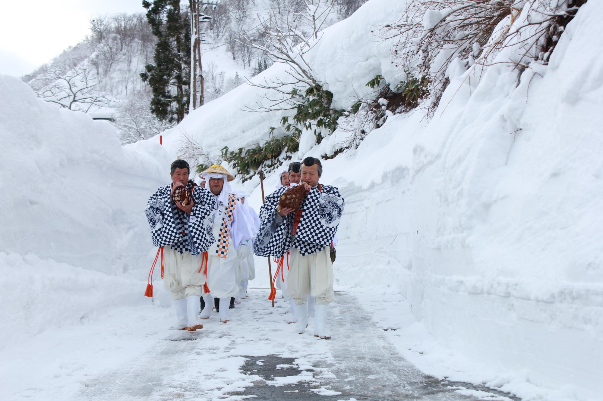 両脇に2メートルほどの雪壁の中、白装束に身を包んだ10人ほどの男性がほら貝をふきながら歩く様子。