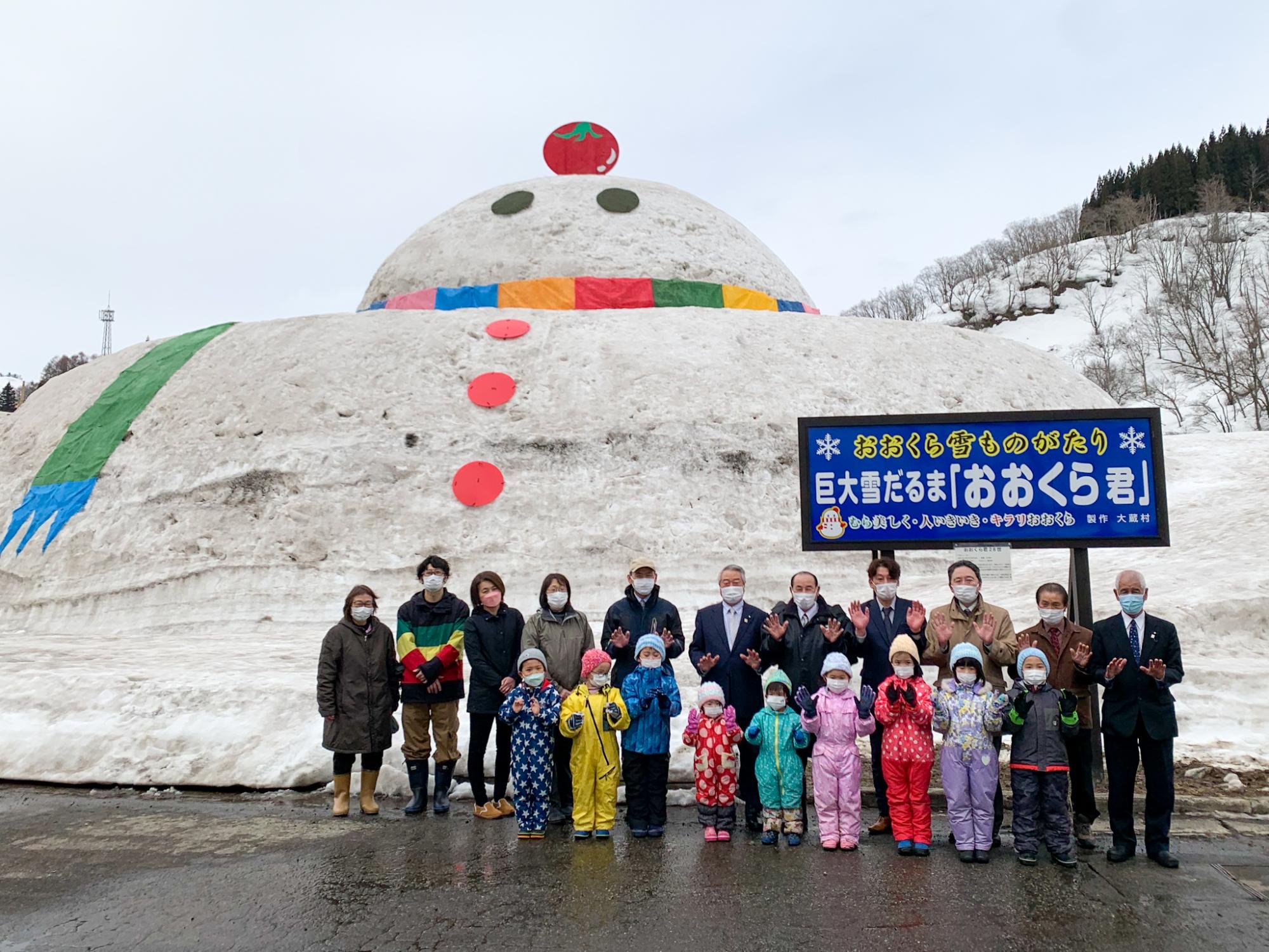 巨大雪だるまの前に防寒着を着て立つ保育所の子どもたちと大人たち