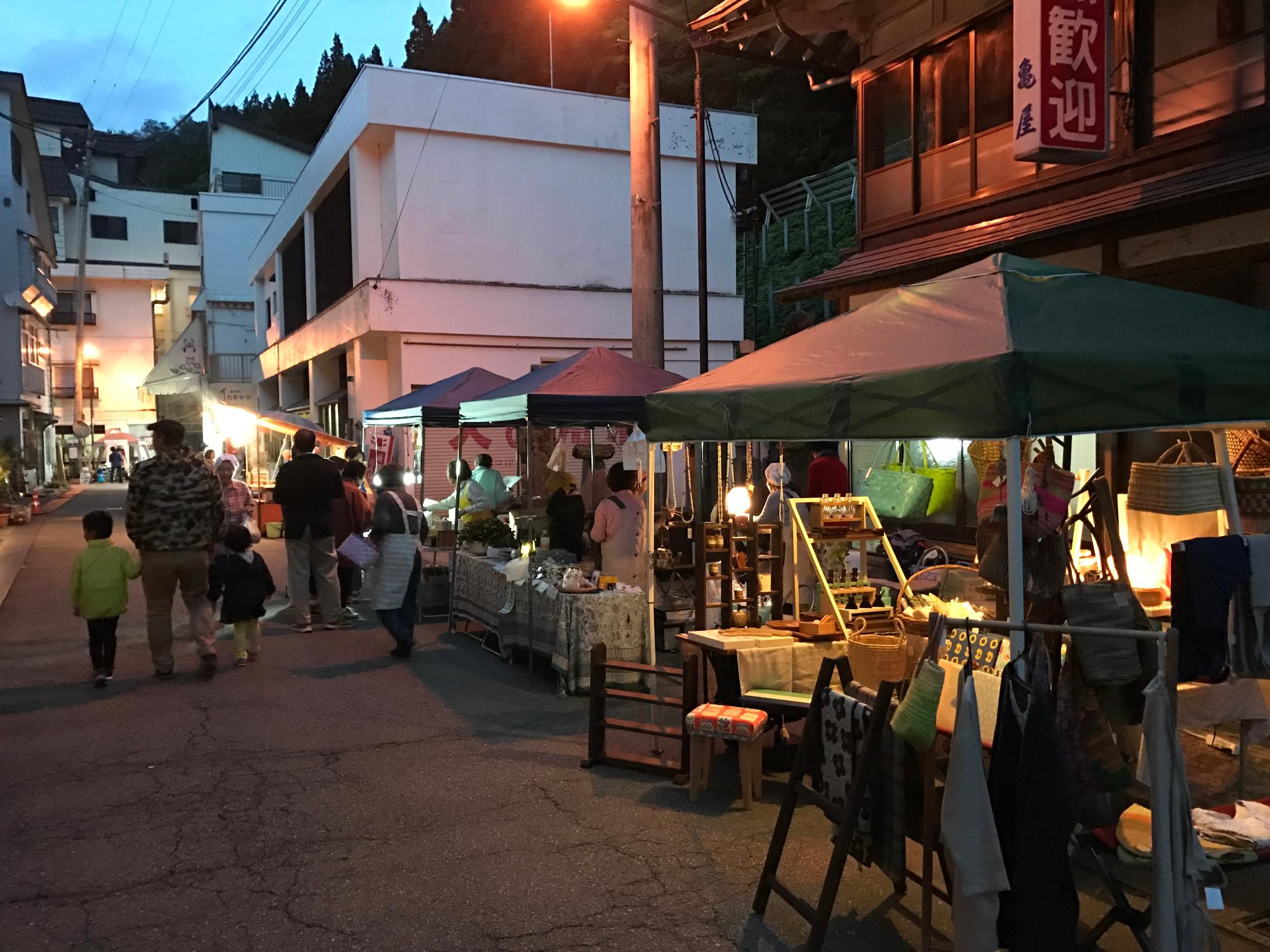 夕暮れの温泉街で旅館の軒先に出店が立ち並び、買い物をする人々の画像